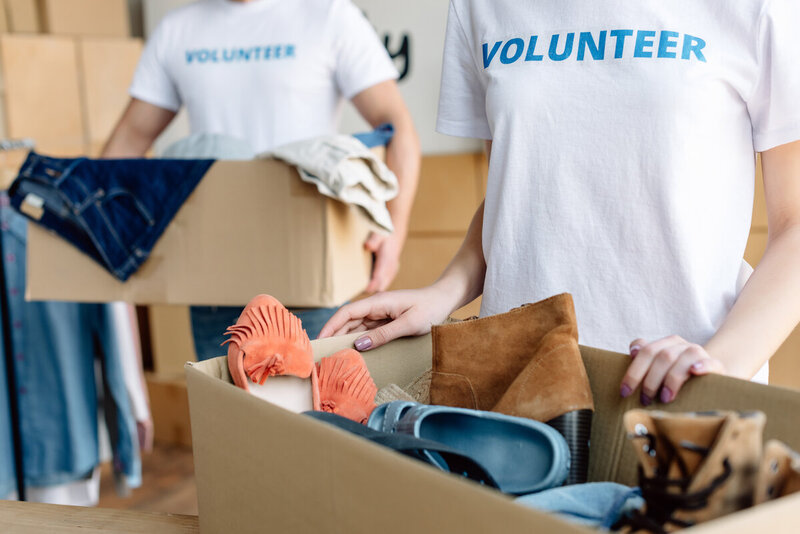 A young volunteer holds a box of donated clothes, while wearing a white tshirt that says volunteer in blue printing.