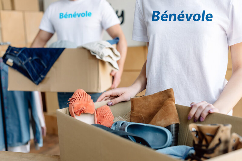 A young volunteer holds a box of donated clothes, while wearing a white tshirt that says volunteer in blue printing.